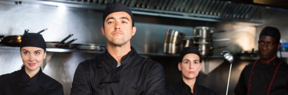 Dois homens e duas mulheres, todos cozinheiros vestidos com uniformes pretos, atrás de uma bancada de restaurante e segurando alguns equipamentos de cozinha.