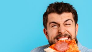 Homem sorridente comendo uma fatia de pizza, fundo da imagem em cor turquesa.
