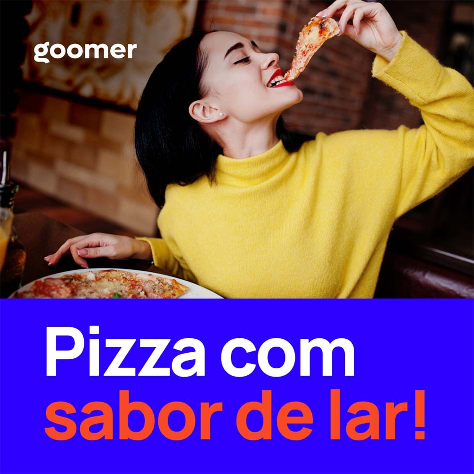 Mulher sentada em uma mesa de pizzaria e comendo uma fatia com uma das mãos, a outra mão apoiada sobre a mesa.