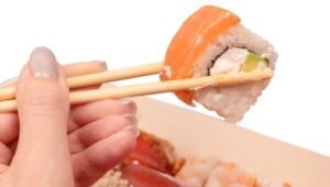 Mão feminina utilizando hashi para segurar uma peça de sushi, fundo da imagem branco