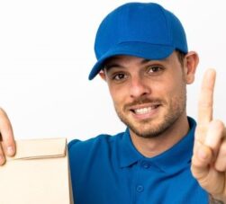 Entregador de delivery vestido com camiseta e boné azul, segurando um pacote de entrega com uma das mãos, e a outra mão sinalizando com o indicador para cima, fundo da imagem branco