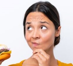 Mulher segurando um hambúrguer com sua mão direita e com a outra mão em seu queixo, com uma expressão de quem está pensando, fundo da imagem branco