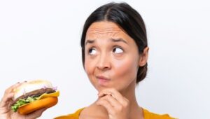 Mulher segurando um hambúrguer com sua mão direita e com a outra mão em seu queixo, com uma expressão de quem está pensando, fundo da imagem branco