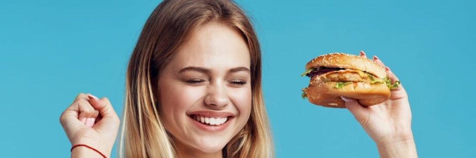Mulher com hambúrguer em uma das mãos com expressão de felicidade, fundo da imagem em azul-claro